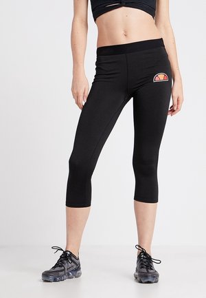 Femme portant un legging noir court et des chaussures de sport noires, debout les jambes écartées sur un fond clair uni.