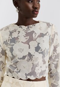 Csipkés crop top virágmintával, krémszínű, áttetsző alapon, hullámos nyakkivágással és hosszú ujjakkal, hozzáillő aljjal párosítva.