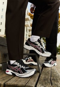 Black and silver athletic sneakers with red accents, featuring a breathable mesh upper and cushioned sole, positioned on a wooden bench.