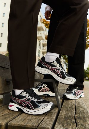 Zapatillas atléticas en negro y plata con detalles en rojo, con parte superior de malla transpirable y suela acolchada, colocadas sobre un banco de madera.