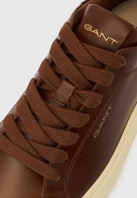 Baskets en cuir marron avec lacets tressés marron, arborant un logo doré "GANT" sur le côté et une semelle en caoutchouc de couleur crème.