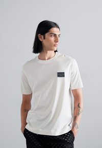 White short-sleeve t-shirt with a round neck and a black label on the left chest; smooth fabric and relaxed fit. Paired with patterned black pants.