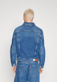 Keskitummansininen farkkutakki, jossa on klassinen leikkaus, nappisuljenta, kaksi sivutaskua ja hillitty logodetalji takana.