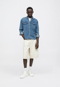 Chemise en denim avec deux poches poitrine, manches retroussées, associée à un short beige clair jusqu'aux genoux et des baskets blanches avec des chaussettes de cheville.