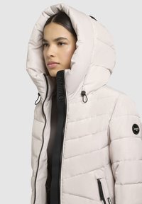 Veste rose pâle rembourrée avec une grande capuche, fermeture éclair devant et détails noirs. Présente un design matelassé et une patch logo sur la manche.