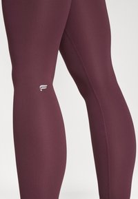 Μπορντό κολάν φτιαγμένα από ελαστικό ύφασμα, με απαλό τελείωμα, εφαρμοστό σχέδιο και μικρό λευκό λογότυπο κοντά στο τελείωμα.