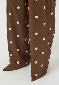 Brune, vide bukser med store hvite polkadotter. Laget av glatt materiale med spisse brune hæler delvis synlige nederst.