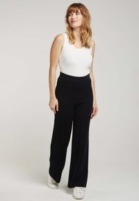 Pantalon large noir avec une taille haute ajustée, associé à un débardeur blanc. Le modèle porte des baskets blanches, affichant un look décontracté.