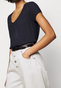 Navy blue short-sleeve V-neck top paired with light-colored, high-waisted trousers featuring large buttons and pocket details.
