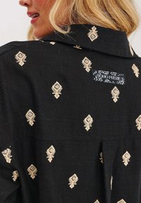 Černá košile s krémovým geometrickým vyšíváním, límečkem a štítkem "BOHO PRERIE" na zadní straně. Měkká látka s texturovaným povrchem.