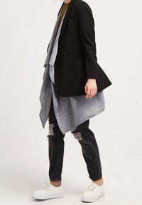 Blazer negro a medida con bufanda asimétrica gris, combinado con jeans negros desgastados y zapatillas blancas. Tejido suave con un diseño estructurado.