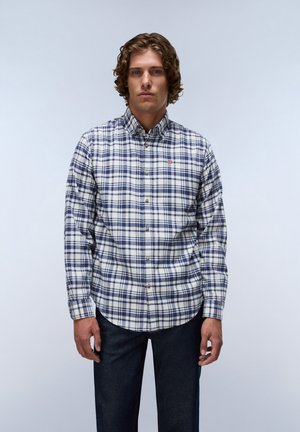 Mand med krøllet hår iført en blå og hvid ternet skjorte og mørke jeans, stående mod en ensfarvet lys baggrund.