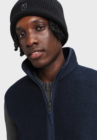 Gilet in pile blu navy con colletto alto, chiusura zip e maglione grigio a coste sotto. Berretto lavorato a maglia nero con dettagli del logo.