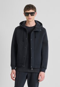 Donker marineblauwe hoodie met een rits aan de voorkant, drukknoopjes, textuurstof en geribde manchetten. Gecombineerd met een zwart shirt eronder.