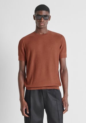 Uomo con occhiali da sole neri, maglione a coste a maniche corte color ruggine e pantaloni neri larghi, in piedi davanti a uno sfondo semplice.