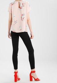 Blouse florale rose clair à manches courtes avec un détail en trou de serrure dans le dos, associée à un legging noir ajusté et des sandales à talons hauts à lanières rouges.