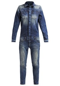 Combinaison en denim à manches longues, boutonnée à l'avant, poches poitrine et détails délavés et usés sur fond blanc.