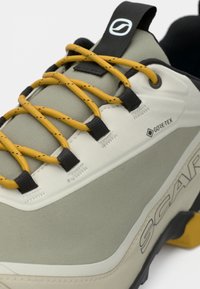 Zapato gris claro y beige con superficie texturizada, cordones amarillos, acentos negros y un logo de GORE-TEX, con una suela de goma resistente.