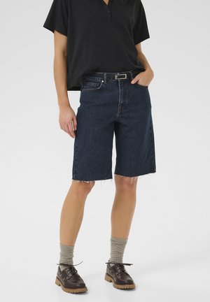 Dunkelblaue Denim-Shorts mit fransigem Saum, kombiniert mit einem schwarzen Poloshirt und braunen Schuhen. Graue Socken sind oberhalb der Schuhlinie sichtbar.