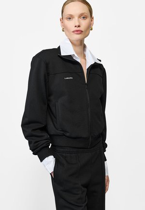 Czarna kurtka na zamek o gładkiej fakturze, z wysokim kołnierzem, kieszenią na przodzie oraz logo "LAMANIA" na klatce piersiowej. Widać białe mankiety koszuli.