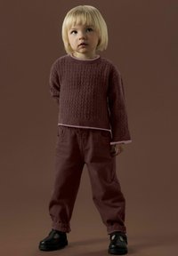 Pull en maille bordeaux avec motif texturé, superposé à un col rose clair ; pantalon bordeaux ample ; chaussures noires. Un enfant se tient debout, les mains derrière le dos.