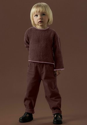 Pull en maille bordeaux avec motif texturé, superposé à un col rose clair ; pantalon bordeaux ample ; chaussures noires. Un enfant se tient debout, les mains derrière le dos.
