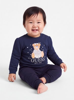 Bébé souriant portant une tenue bleu marine avec un motif d'ours en peluche et "GUESS EST. 1981", assis pieds nus sur un fond blanc.