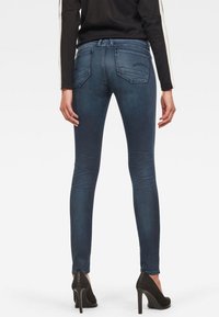 Jeans ajustados de mezclilla oscura con una textura suave y diseñados con bolsillos traseros. Llevados con zapatos de tacón negros, vistos desde atrás.