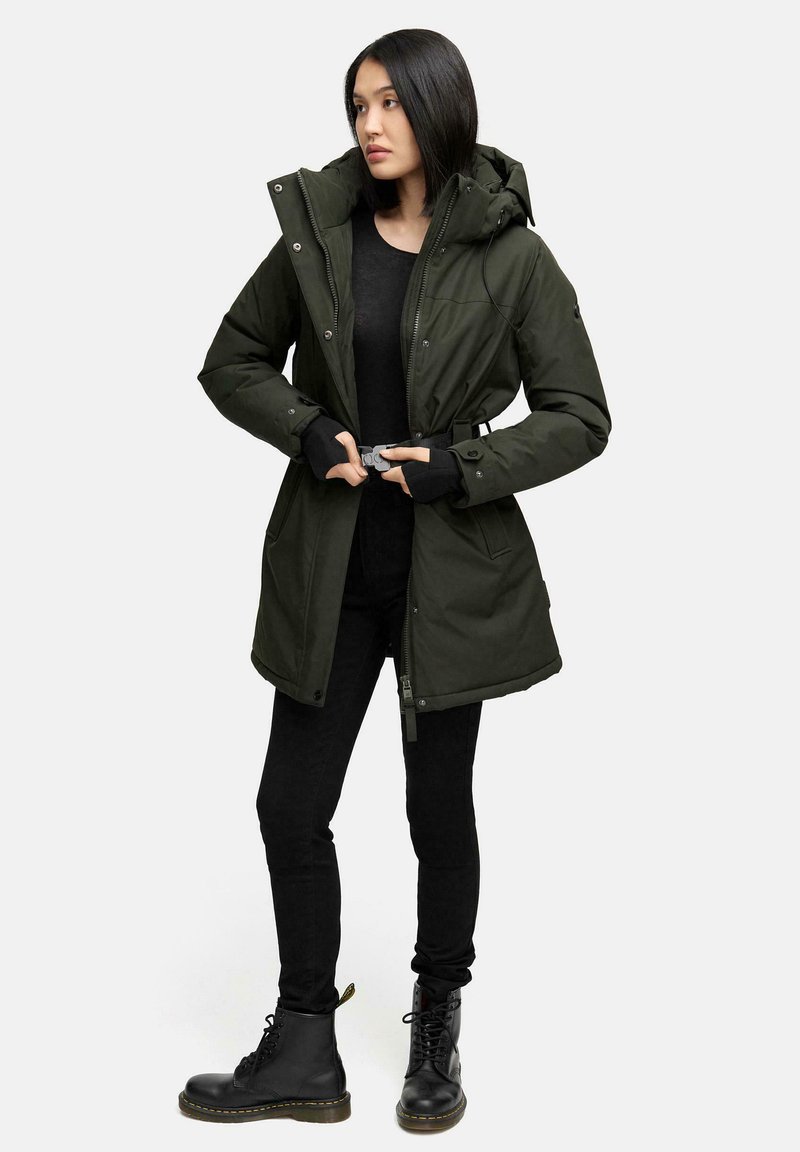 Parka verte matelassée avec une capuche, fermeture éclair et pression, poches latérales et taille ajustable. Portée sur un haut noir et un pantalon noir avec des bottes.
