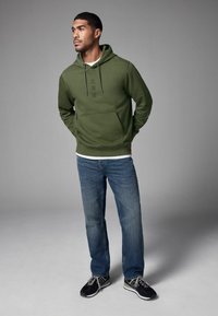Sweatshirt vert avec une poche avant et des lettres noires, associé à un jean bleu et des chaussures de sport noires. Design simple et décontracté.