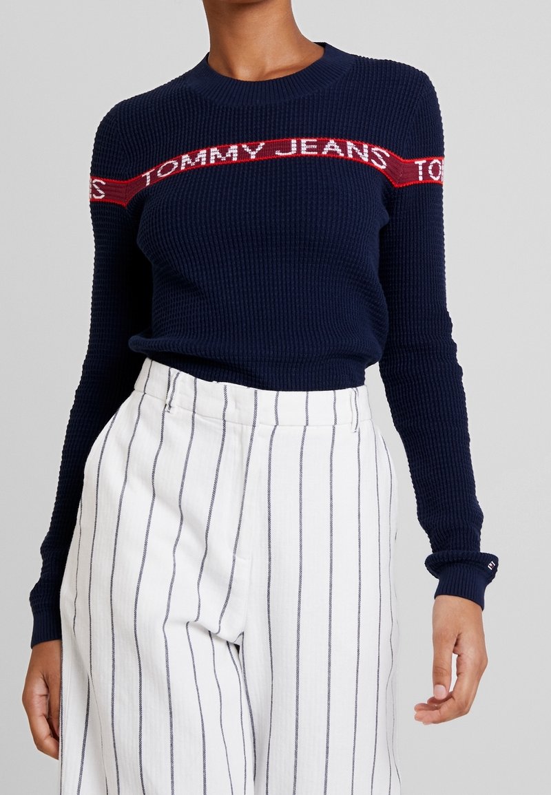Maglione a coste blu navy con striscia rossa con logo "Tommy Jeans" sul petto, abbinato a pantaloni bianchi a vita alta con sottili strisce verticali.