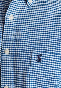 Niebiesko-biała koszula w kratę z białymi guzikami i małym granatowym haftowanym ptakiem na kieszeni piersiowej.