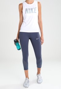 Valkoinen hihaton Nike-toppu, jossa on heijastava logo, yhdistettynä tummansinisiin capri-leggingseihin ja vaaleanharmaisiin urheilukenkät. Pitelee turkoosia vesipulloa.