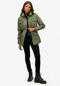 Chaqueta de utilitario verde oliva con dos bolsillos en el pecho, cierre de cremallera y hombreras, usada sobre una blusa negra y pantalones negros, combinada con botas negras.