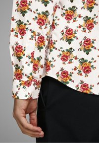 Koszula w białej bawełnie z kwiatowym wzorem, czerwonymi i żółtymi różami oraz zielonymi liśćmi, z guzikami na mankietach. Gładka faktura, zaokrąglony dół.