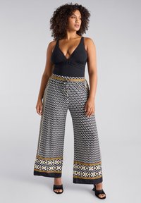 Zwarte V-hals bodysuit met een knoopdetail, gecombineerd met wijde broekspijpen met een zwart-wit hartpatroon en een contrasterende gouden accentband rond de taille.