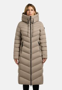 Donna che indossa un lungo cappotto imbottito beige a righe con colletto alto e cerniera nera, in piedi davanti a uno sfondo bianco semplice.