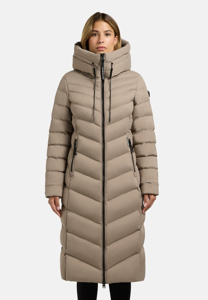 Donna che indossa un lungo cappotto imbottito beige a righe con colletto alto e cerniera nera, in piedi davanti a uno sfondo bianco semplice.