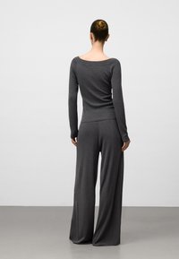 Kvinna med håret i en knut, iklädd en figurnära mörkgrå långärmad topp och vida byxor, stående mot en enkel ljus bakgrund.