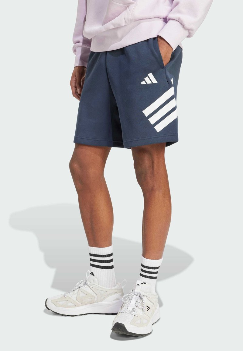 Navy cotton shorts with white three-stripe logo design, elastic waistband, and side pockets. Paired with white sneakers and black-striped socks.