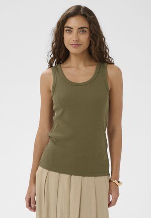 Femme aux cheveux bouclés portant un haut sans manches côtelé vert olive et une jupe plissée beige, debout devant un fond uni.