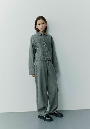 Graue Jeansjacke mit zwei Taschen und Knöpfen, kombiniert mit lockeren grauen Jeanshosen. Schwarze klobige Schuhe vervollständigen das Outfit, vor einem schlichten Hintergrund.