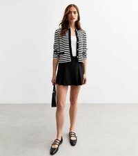 Cardigan a strisce bianche e nere con bottoni dorati, abbinato a una gonna nera a pieghe e scarpe flat nere. Gli accessori includono una piccola borsa.