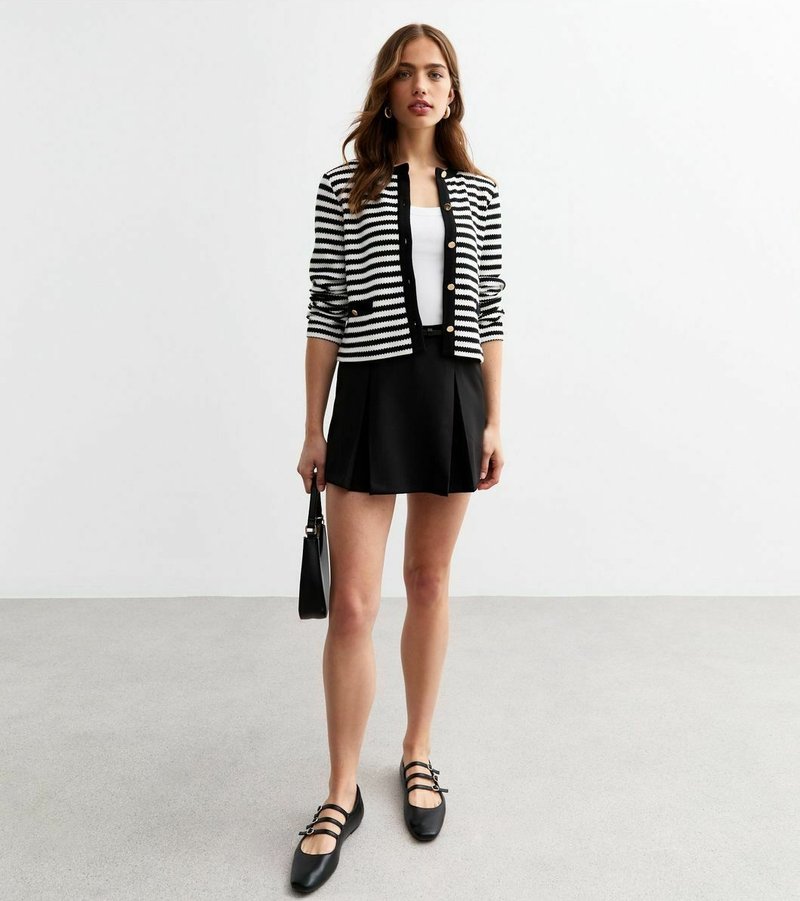 Cardigan a strisce bianche e nere con bottoni dorati, abbinato a una gonna nera a pieghe e scarpe flat nere. Gli accessori includono una piccola borsa.