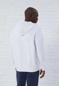 White hooded sweatshirt with a relaxed fit, soft texture, and ribbed cuffs, paired with dark blue pants. Background shows a white brick wall.