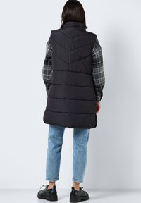 Svart puffervest med chevron-quiltet tekstur, høy krage og ermeløs design, sammen med blå oppbrettede jeans og svarte sko.