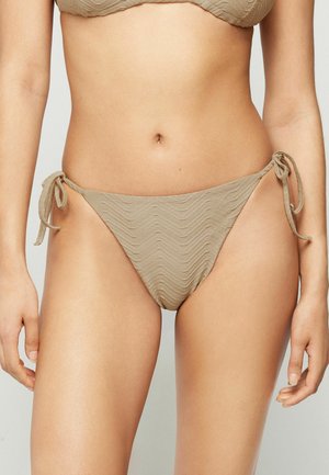 Kobieta w beżowym, teksturowanym bikini z wiązaniami po bokach, pokazująca tors i górną część ud na jednolitym tle.