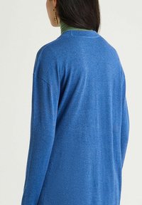 Pull bleu à coupe décontractée, manches longues et encolure ronde. La texture semble douce, avec une légère brillance et un design léger.