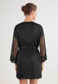 Robe nero in satin con maniche lunghe in pizzo trasparente a pois e orlo in pizzo. Dettaglio in vita annodato e texture liscia, che offre una silhouette fluida.