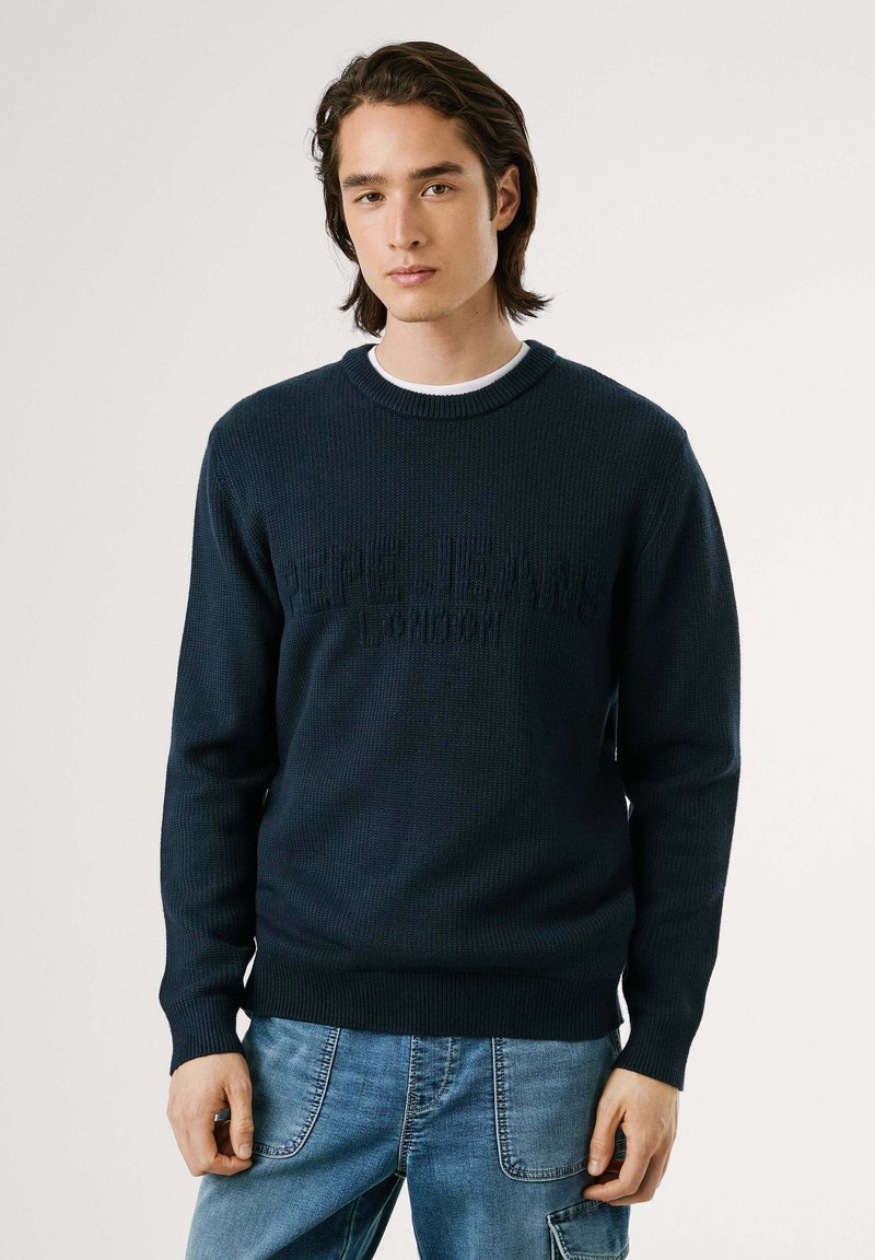 Tamsiai mėlynas megztinis su raišteliais ir apačia, turintis tekstūruotą logotipo dizainą. Dėvimas ant balto apvalaus kaklo marškinėlių, derinamas su džinsiniais džinsais.